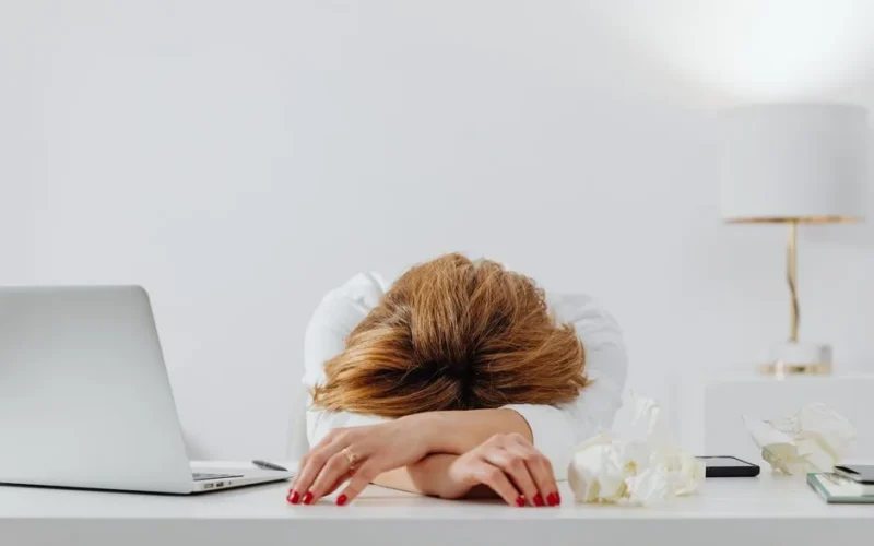 Tired woman resting head on arms at desk with laptop, showing fatigue in modern office.