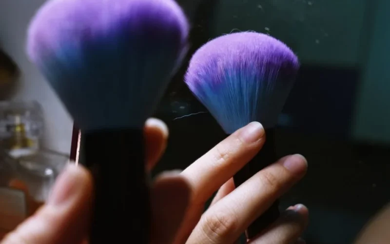 Close-up of a hand holding a purple-bristled makeup brush, reflecting in a mirror.