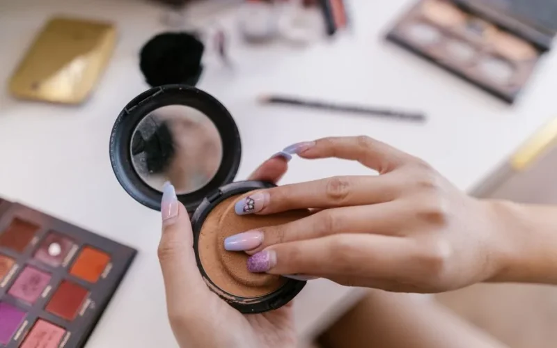 Hands applying powder with a sponge from a compact, surrounded by makeup products.