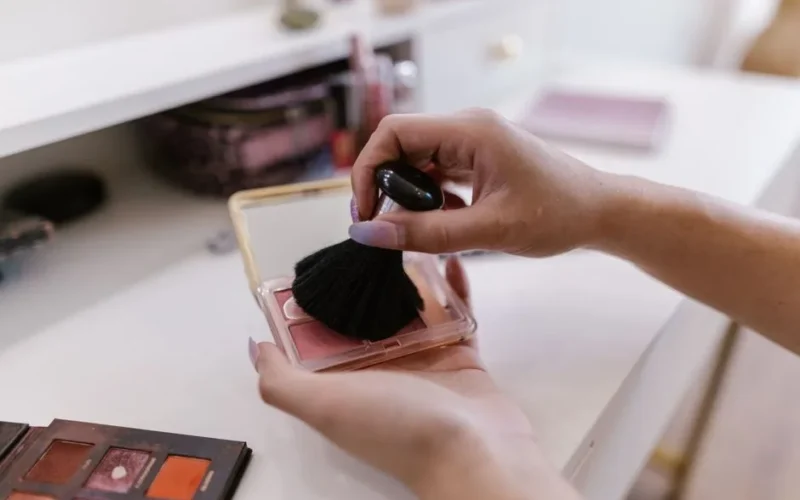 Close-up of woman's hands applying blush with a brush, indoors.