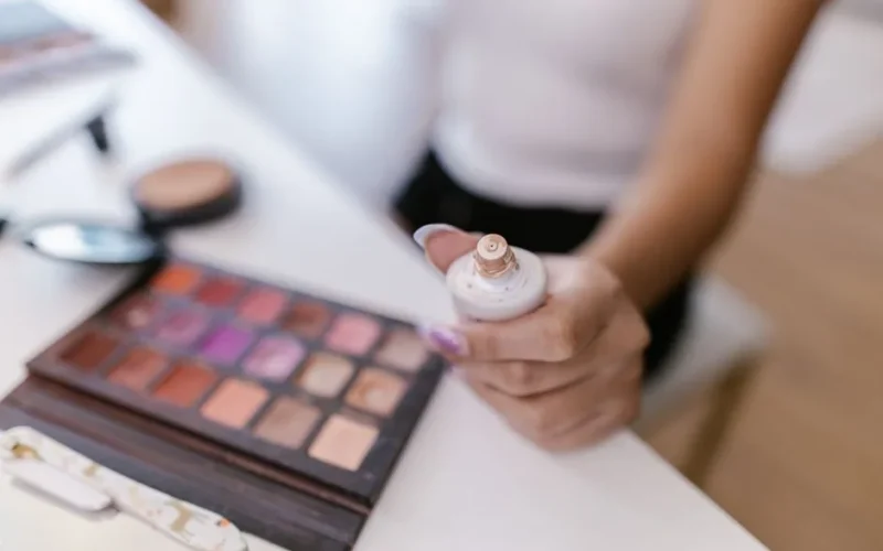 Close-up of a person holding foundation bottle with colorful makeup palette in focus.