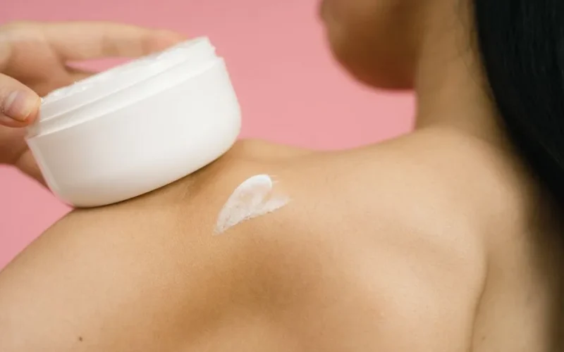 Woman applying skincare cream to shoulder against a pink background.