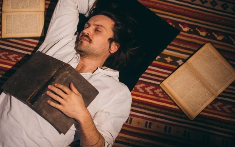 Adult man peacefully sleeping on striped carpet surrounded by open books indoors.