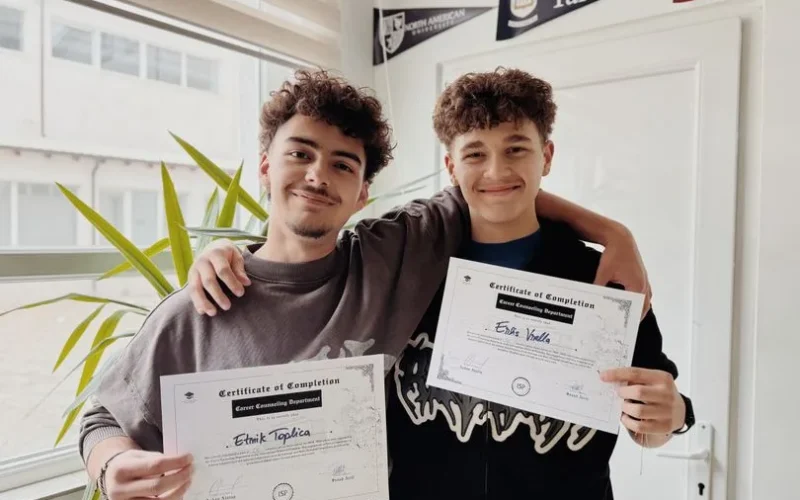 Two young adults proudly display their graduation certificates inside a classroom.