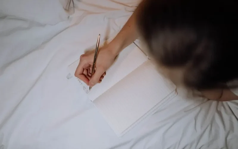 A person writing a to-do list in a notebook while sitting on a bed with white linen.