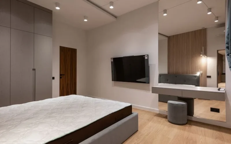 Comfortable bed and wardrobe in modern bedroom placed in front of TV hanging on wall near dressing table with big mirror