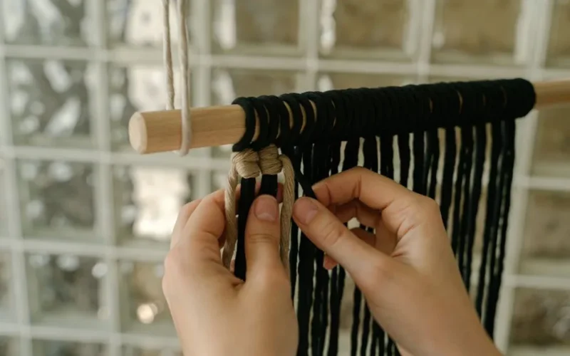 Hands weaving a macrame wall hanging using black and beige yarn. Arts and crafts in focus.