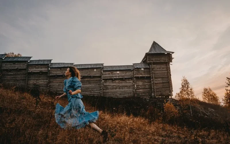 A woman in a blue dress runs by a rustic wooden fence during sunset.