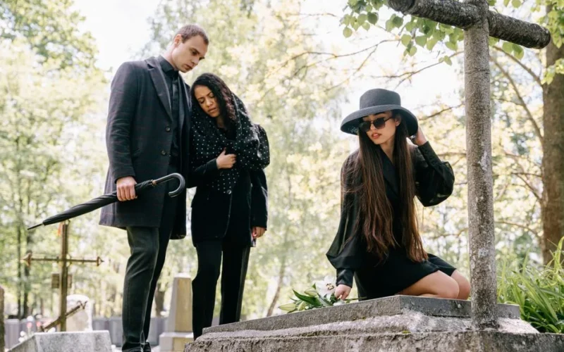 Three adults in a cemetery grieving at a grave during daylight.