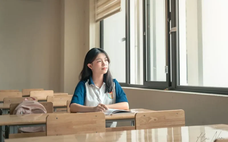 A student sits alone in a classroom, gazing out the window, contemplating. Bright natural light fills the room.