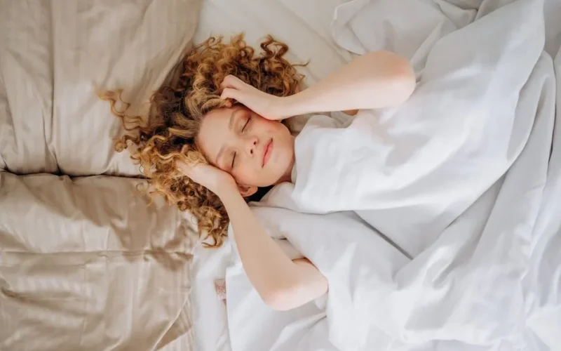 Woman with curly hair sleeping peacefully under white sheets. Indoor comfort.