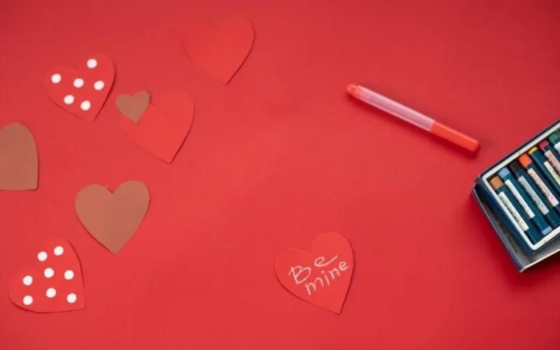 Top view of Be Mine inscription written on paper heart and placed on red background with felt pens during valentines day