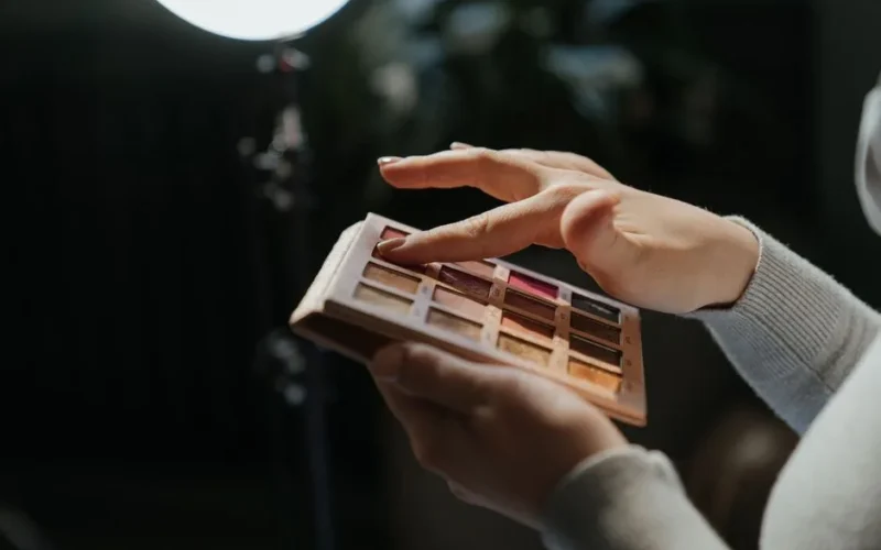 Hands applying eyeshadow from a palette under studio lighting.