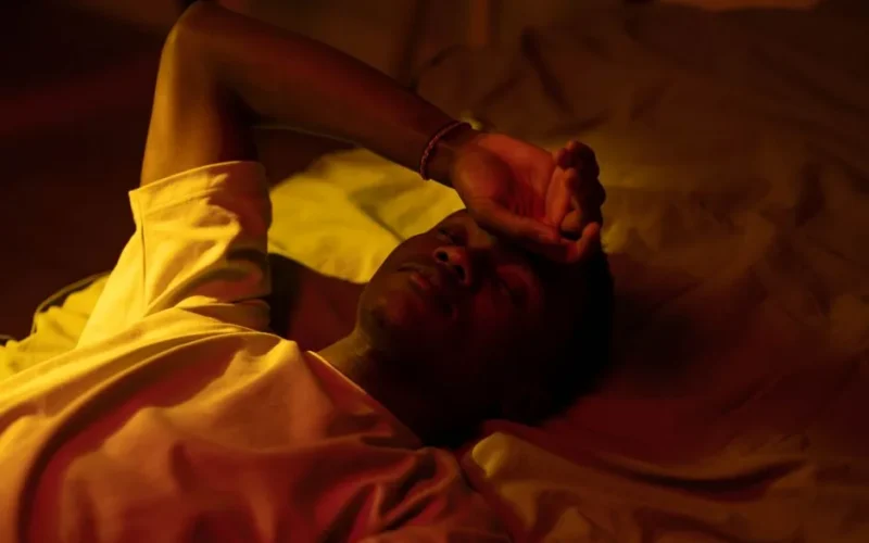 A man lying in bed at night with a moody expression and warm lighting.