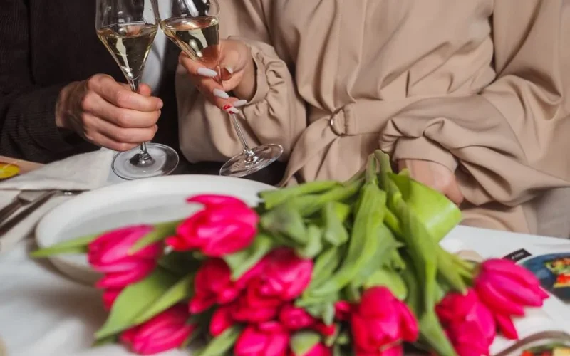 A couple toasting with champagne glasses over a bouquet of pink tulips on a dining table.