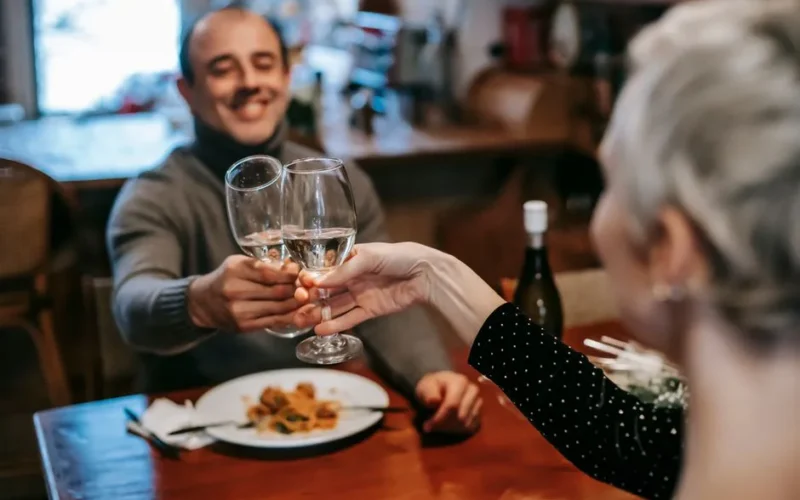 Cheerful adult couple in casual outfits clinking glasses with white wine while having dinner in restaurant