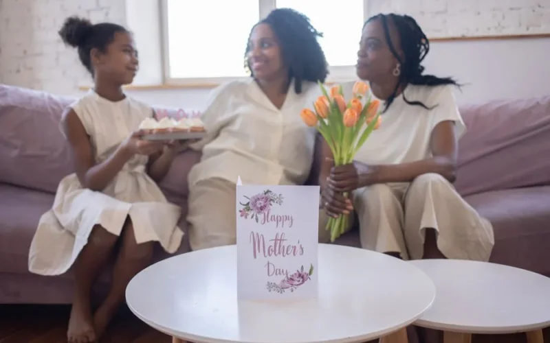 A mother and daughter enjoy a Mother's Day celebration with flowers and cake indoors.