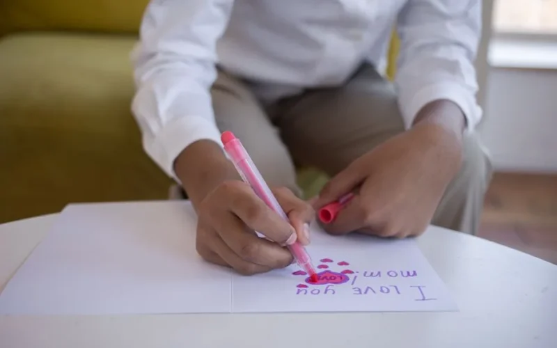 A close-up of a child writing 'I Love You Mom' on a card with a pink marker.