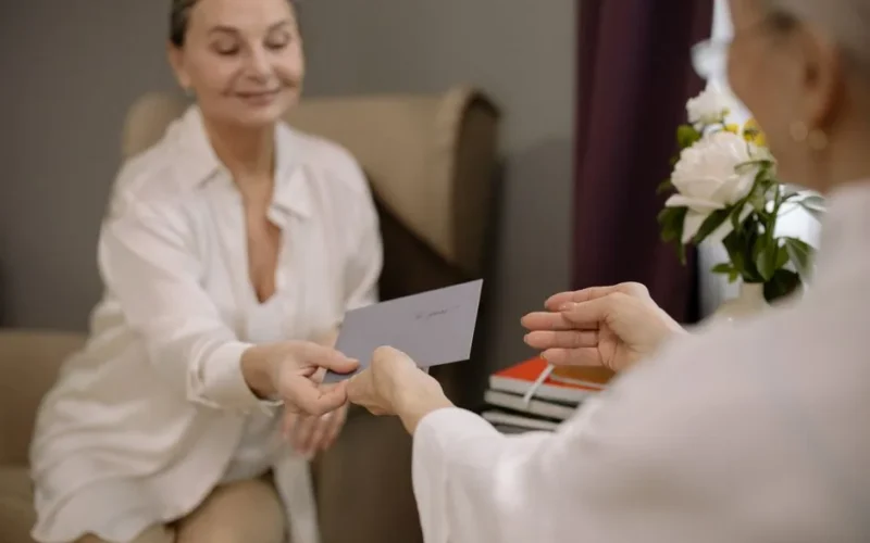 A senior woman passes a greeting card to another person. Warm indoor setting with focused expressions.
