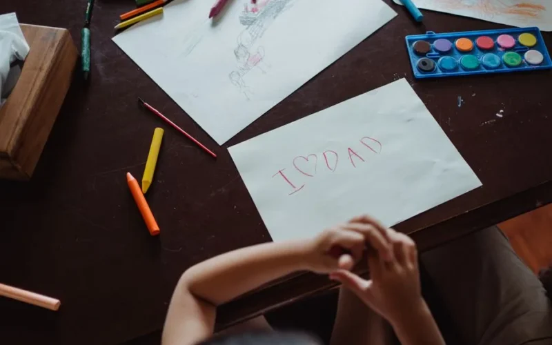 A child's handwritten note 'I ❤️ Dad' on paper surrounded by art supplies on a wooden table.