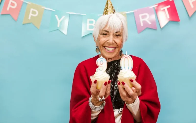 Smiling elderly woman holding cupcakes with candles on 90th birthday party.