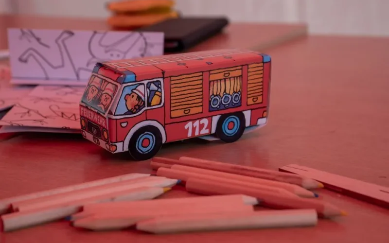 A vibrant paper model fire truck on a table surrounded by colored pencils, perfect for creative activities.