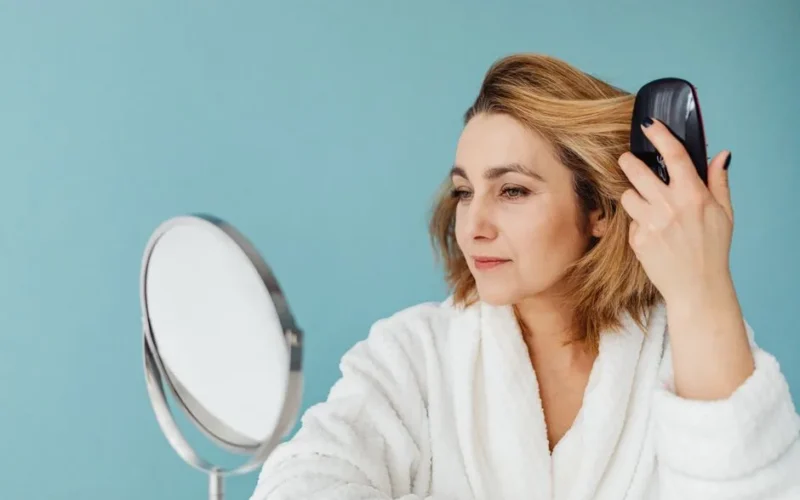 A woman in a bathrobe combs her hair while looking into a mirror with a calm expression.