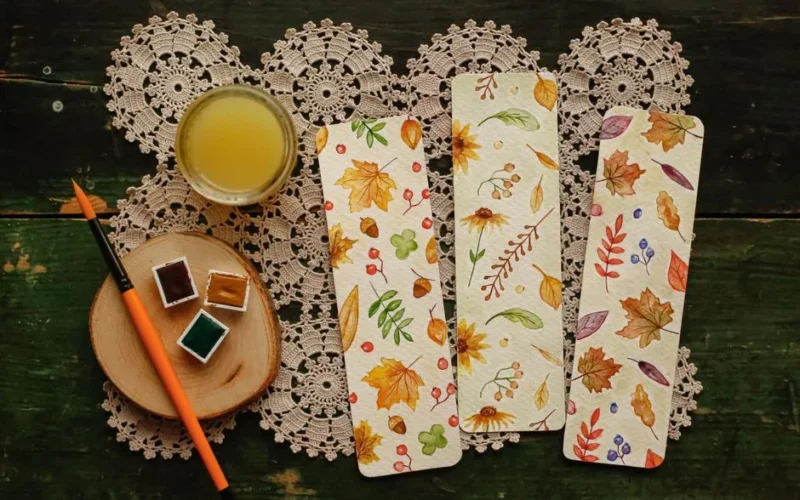 Artistic flat lay featuring watercolor paints, brush, and autumn-themed bookmarks on crochet doily.