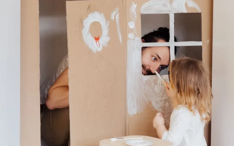 Father and daughter enjoying creative bonding by painting a cardboard playhouse indoors.