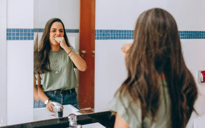 A young woman brushes her teeth, looking in the bathroom mirror. Self-care and hygiene concept.