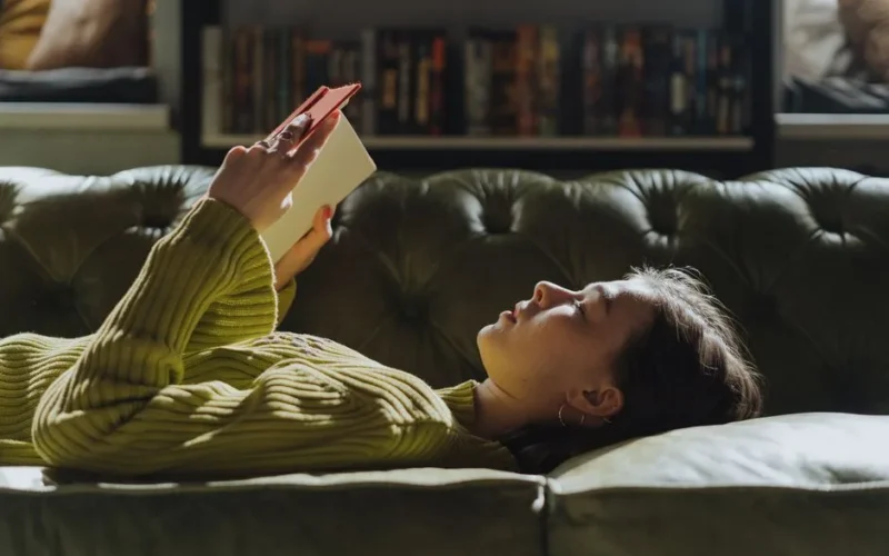A young woman lies on a sofa reading a book, enjoying a peaceful indoor moment.