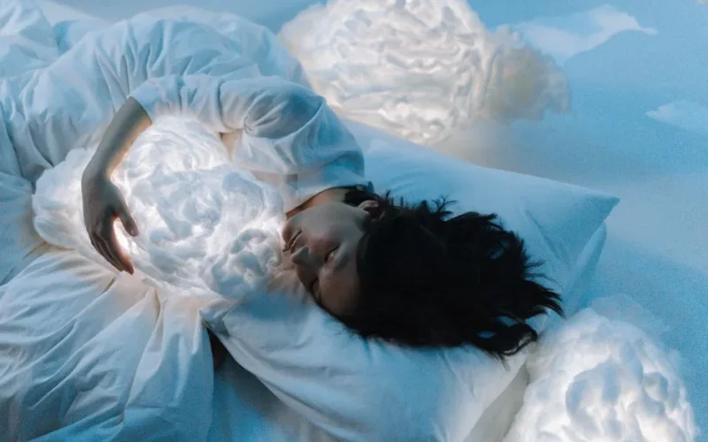 Woman in white hugging glowing cloud pillows in serene, dreamlike setting.