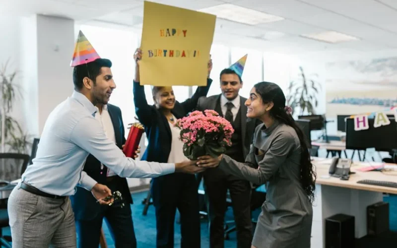 Happy office birthday celebration with colleagues exchanging gifts and wearing party hats.