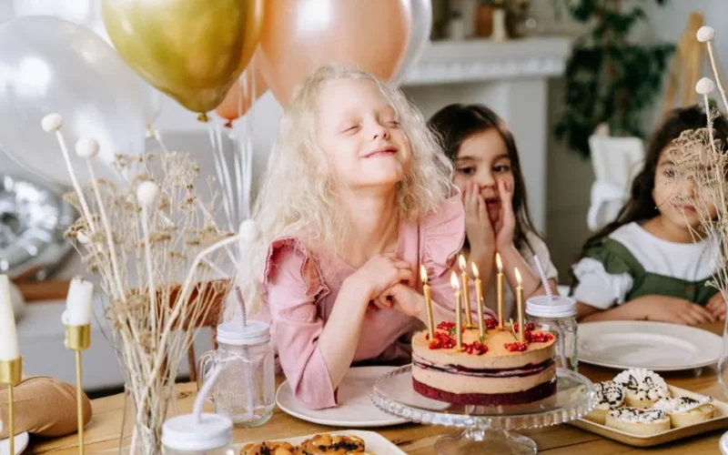 A happy girl makes a wish with lighted candles on her birthday cake, surrounded by friends and balloons.