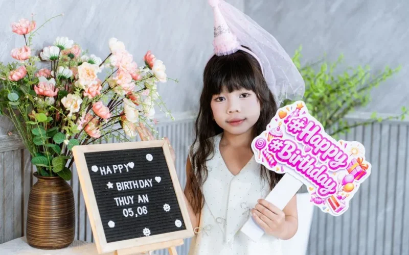 Young girl celebrating birthday indoors with flowers and sign.